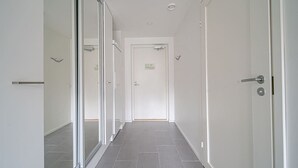Un couloir étroit, carrelé, aux murs et aux portes blancs, qui mène à une pièce dont la porte se trouve au fond.