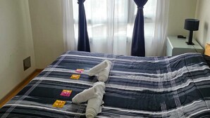 Una cama bien hecha con ropa de cama a rayas y toallas enrolladas, un ventilador de techo y una ventana con cortinas oscuras.