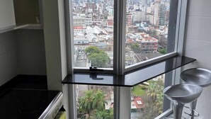 Una cocina en esquina con un ventanal grande, una estufa, un cesto de basura y dos taburetes de bar.