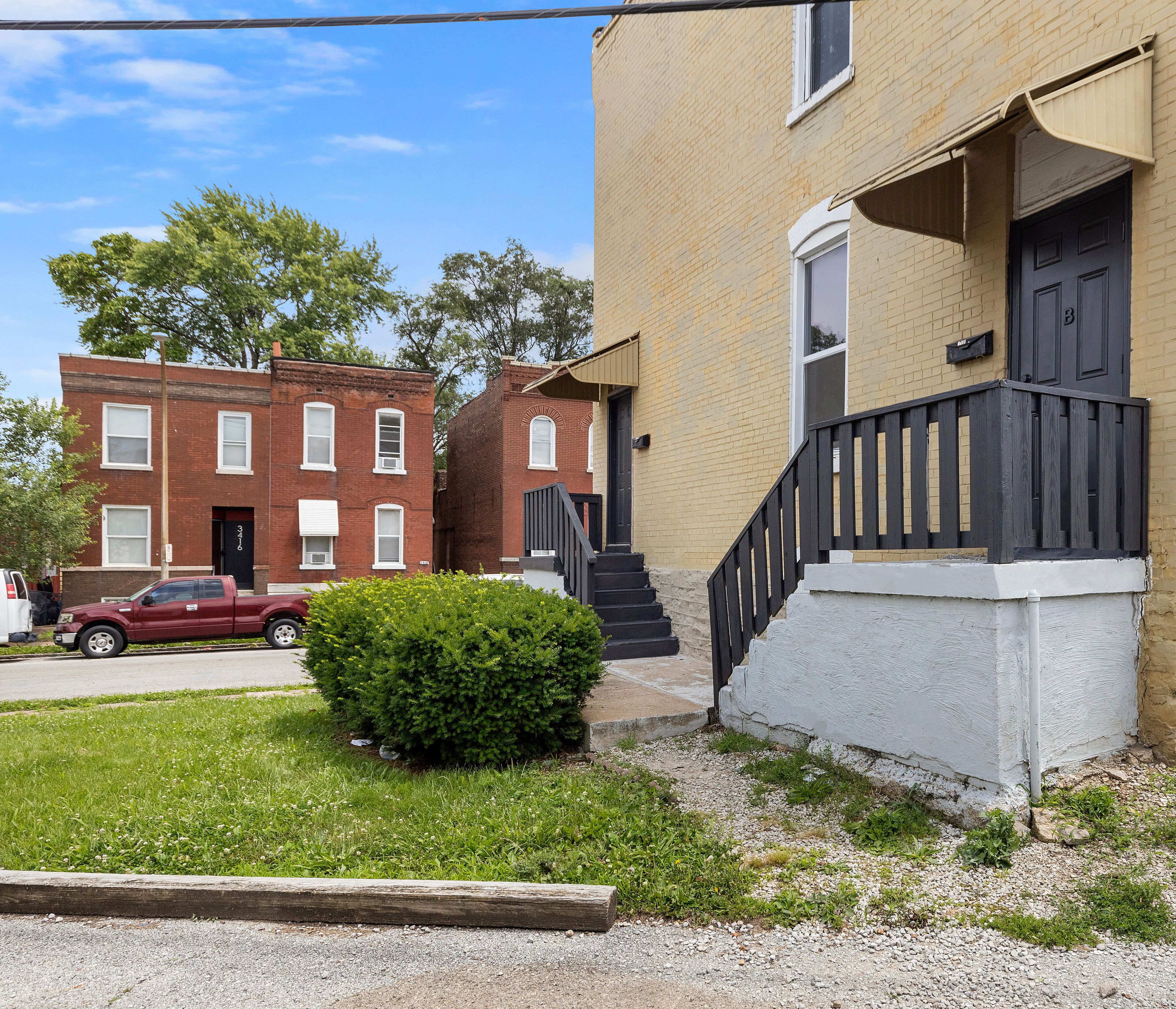 A residential building with a brick facade, a small front yard, and a red car parked on the street.