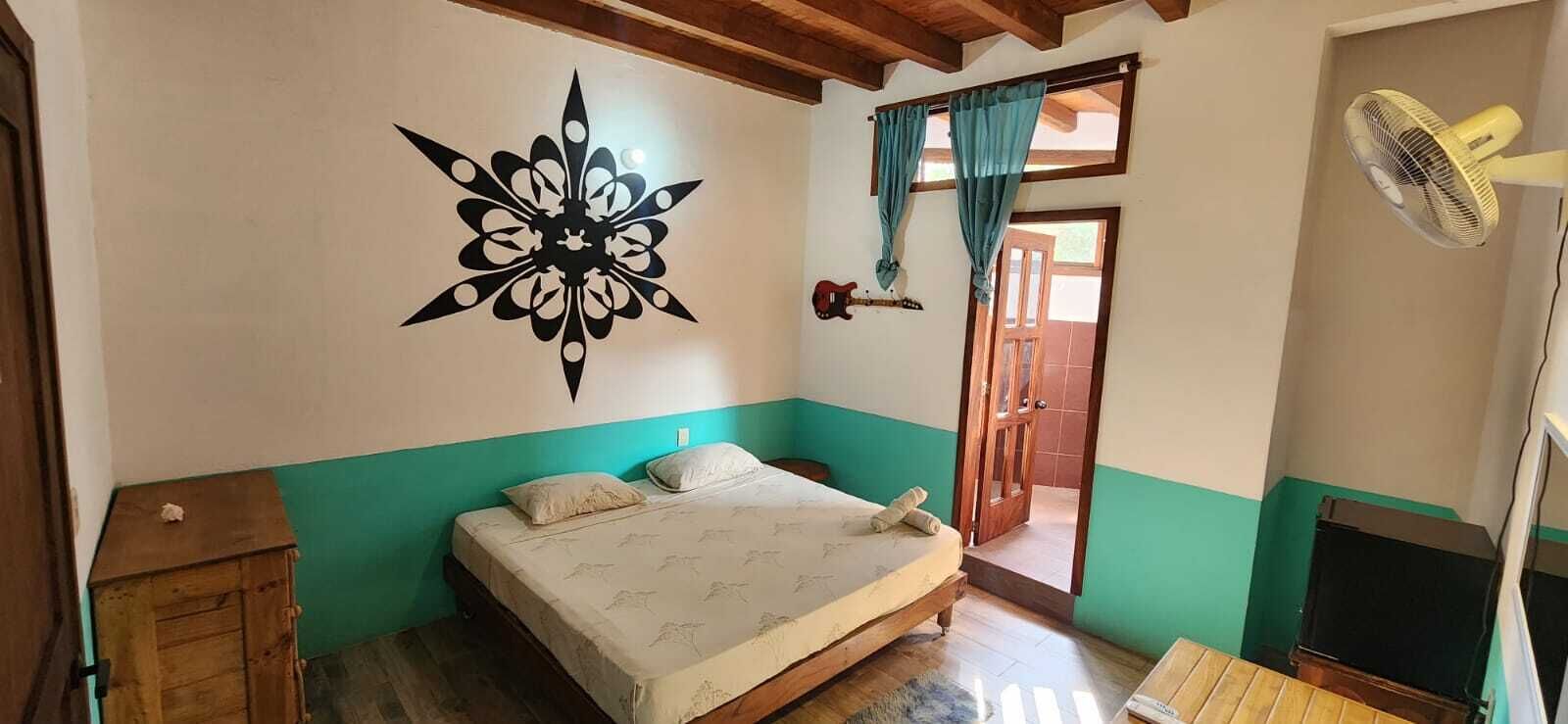 A bedroom with a bed, a wooden cabinet, a decorative wall piece, a guitar, a fan, and a door leading to another room.
