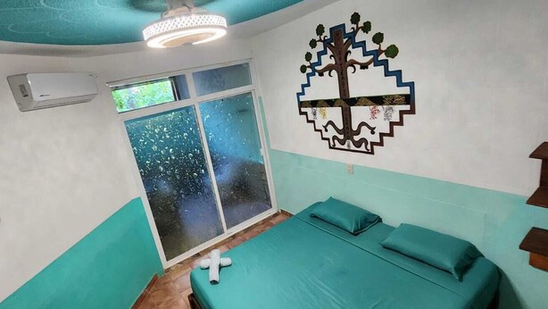 A room with teal walls, a bed with teal bedding, a window with a view of the sky, and a wall decoration of a tree. - La Casa Del Sueño (Mazunte)