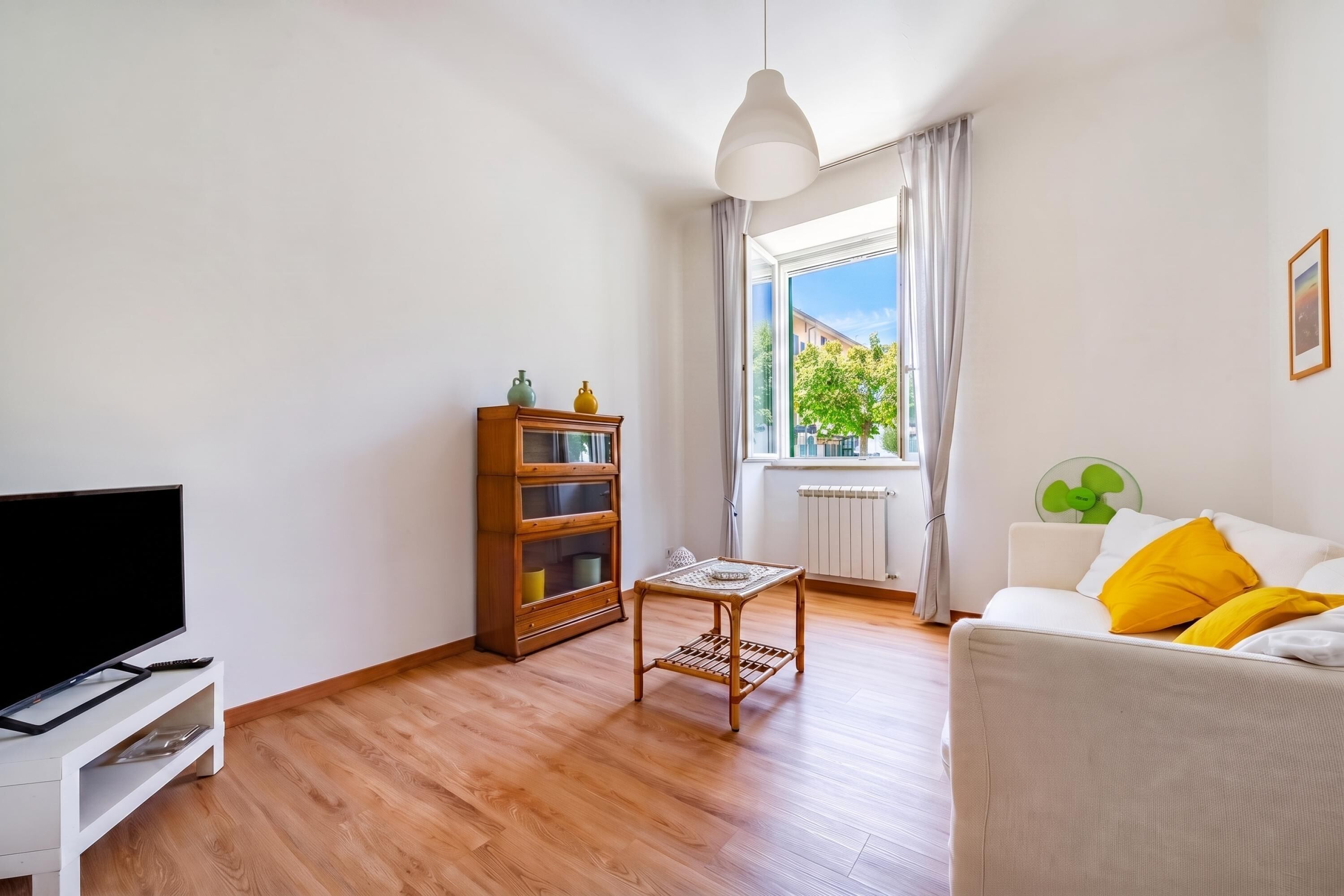 Una sala de estar con televisión de pantalla plana, muebles de madera, un ventanal con cortinas y un ventilador verde.