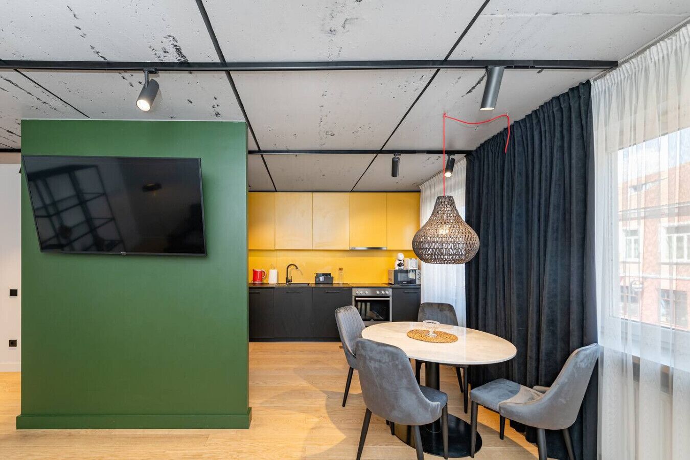 A modern kitchen with yellow cabinets, a dining area with a round table and chairs, and a TV mounted on a green partition.