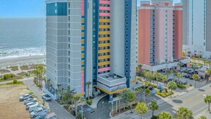 A multi-story hotel with a beach view, featuring a parking lot and a landscaped area with palm trees. - Ocean blue Paradise, Perfect Location. (Myrtle Beach)