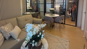 A modern living room with a sofa, a coffee table with a vase of flowers, a chandelier, and a glass partition leading to a kitchen area.