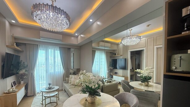 A modern living room with a round marble table, grey upholstered chairs, a chandelier, and a large mirror reflecting the room.
