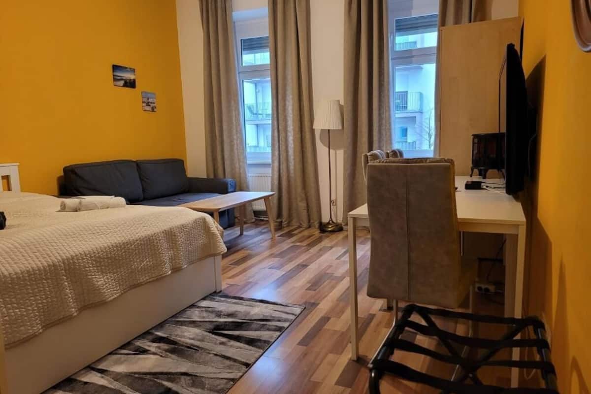 A bedroom with a bed, a desk, a chair, and a view of buildings through large windows.