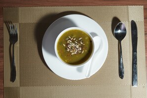 A bowl of soup on a white plate with a spoon and fork beside it, placed on a woven placemat. - Rishikulam Wellness Centre (Samalkha)