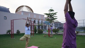 Two people practicing yoga outdoors on pink mats with a playground and a building with a dome in the background.