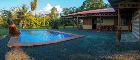 A pool with a wooden border, surrounded by gravel, a wooden structure with a red roof, and a statue of a dog.