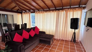 Una sala de estar con techo de madera, un sofá con cojines rojos, un televisor sobre un trípode y piso de baldosas.