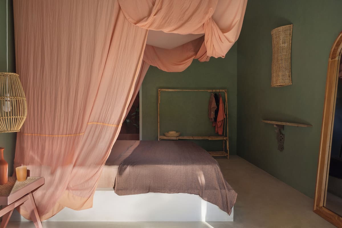 Un dormitorio con cama con dosel, una mesita de noche con un florero, una cesta tejida y un espejo en la pared.