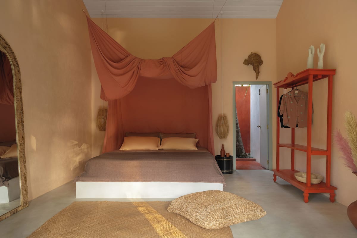 Un dormitorio con una cama grande, un dosel rojo, un espejo, un estante rojo con zapatos y una puerta que da a otra habitación.