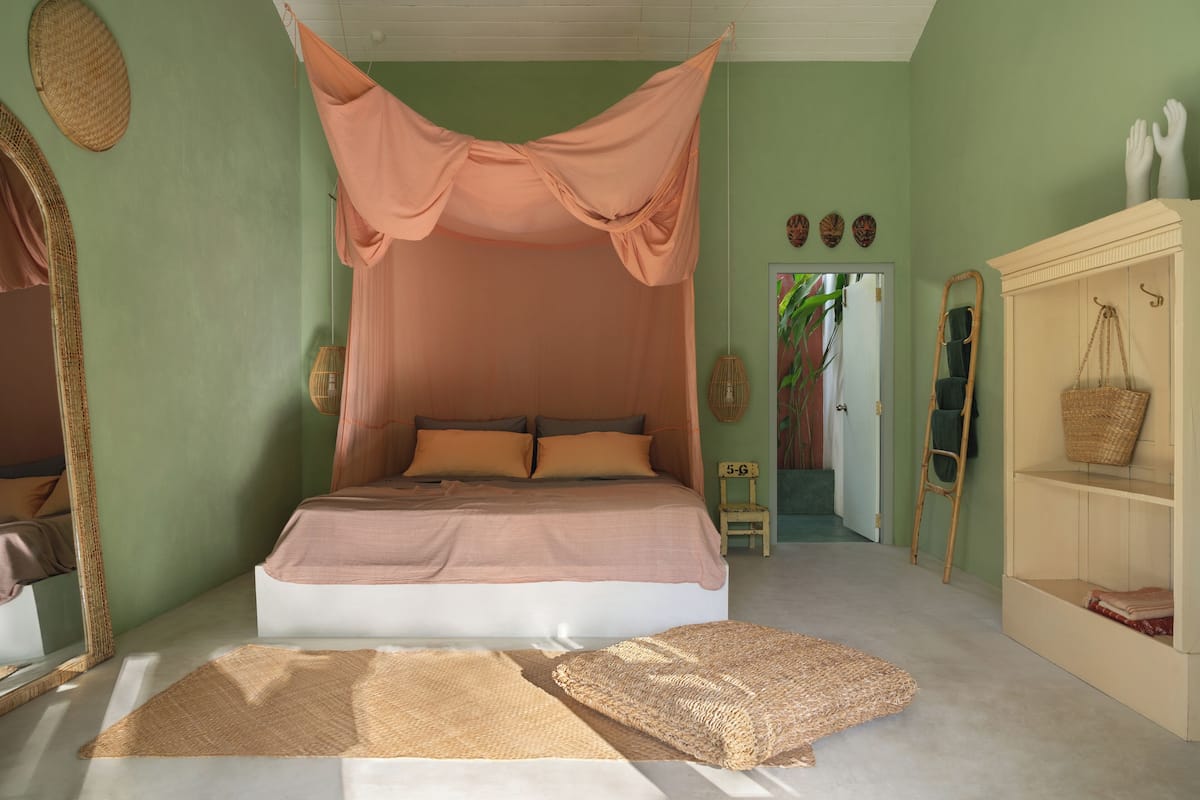 Un dormitorio con cama a dosel, un espejo, una repisa con cestas y una puerta que da a otra habitación.