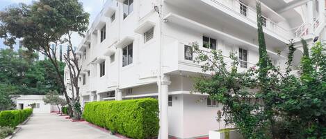 A multi-story white building with balconies, a red entrance, and green landscaping.