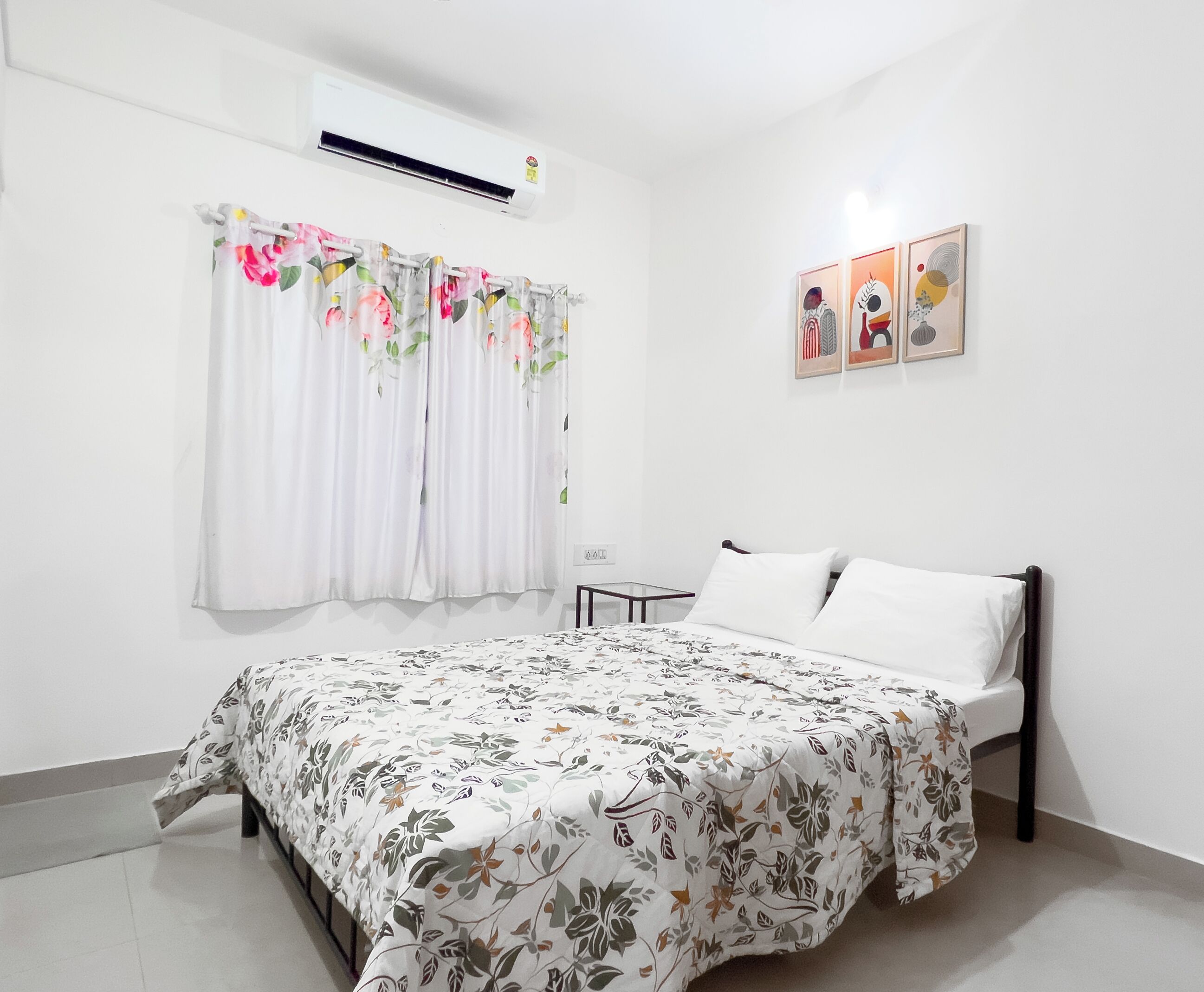 A bedroom with a bed, a window with floral curtains, an air conditioning unit, and three framed artworks on the wall.