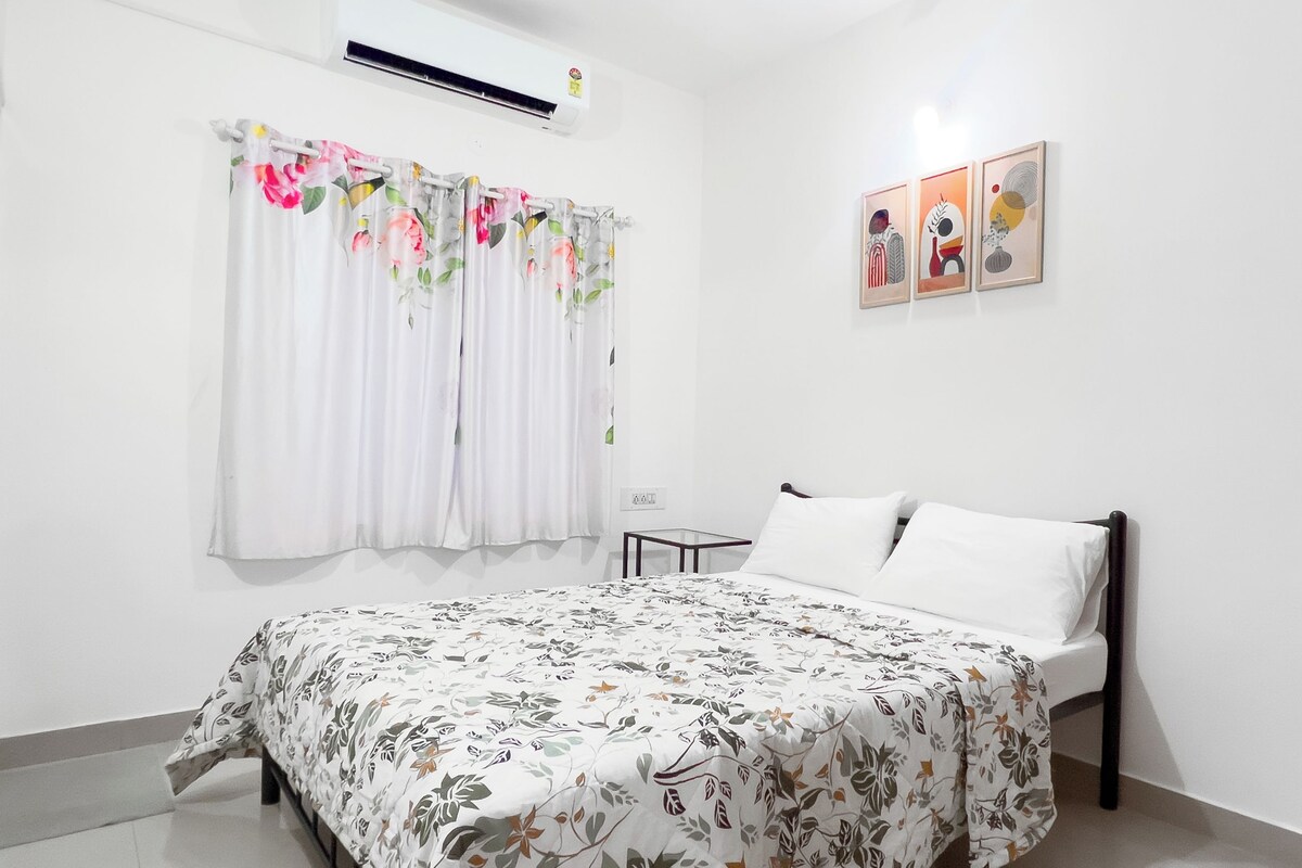 A bedroom with a bed, a window with floral curtains, an air conditioner, and three framed artworks on the wall.
