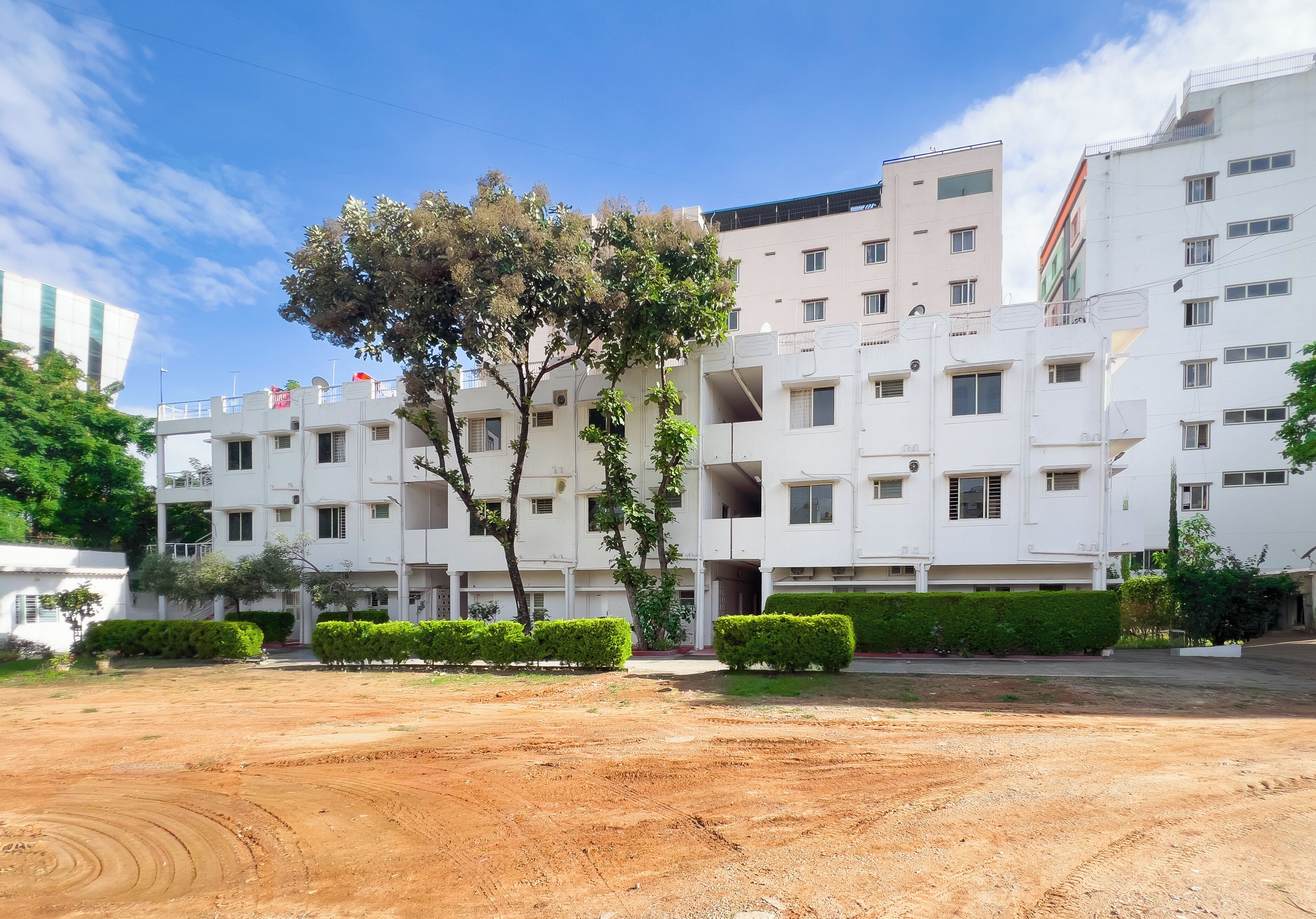 Un edificio blanco de varios pisos con techo plano, rodeado de vegetación cuidada y terreno de tierra.