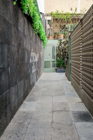 Une ruelle étroite, bordée d’un côté par un mur de pierre et de l’autre par une clôture métallique, qui mène à une entrée fermée par un portail.