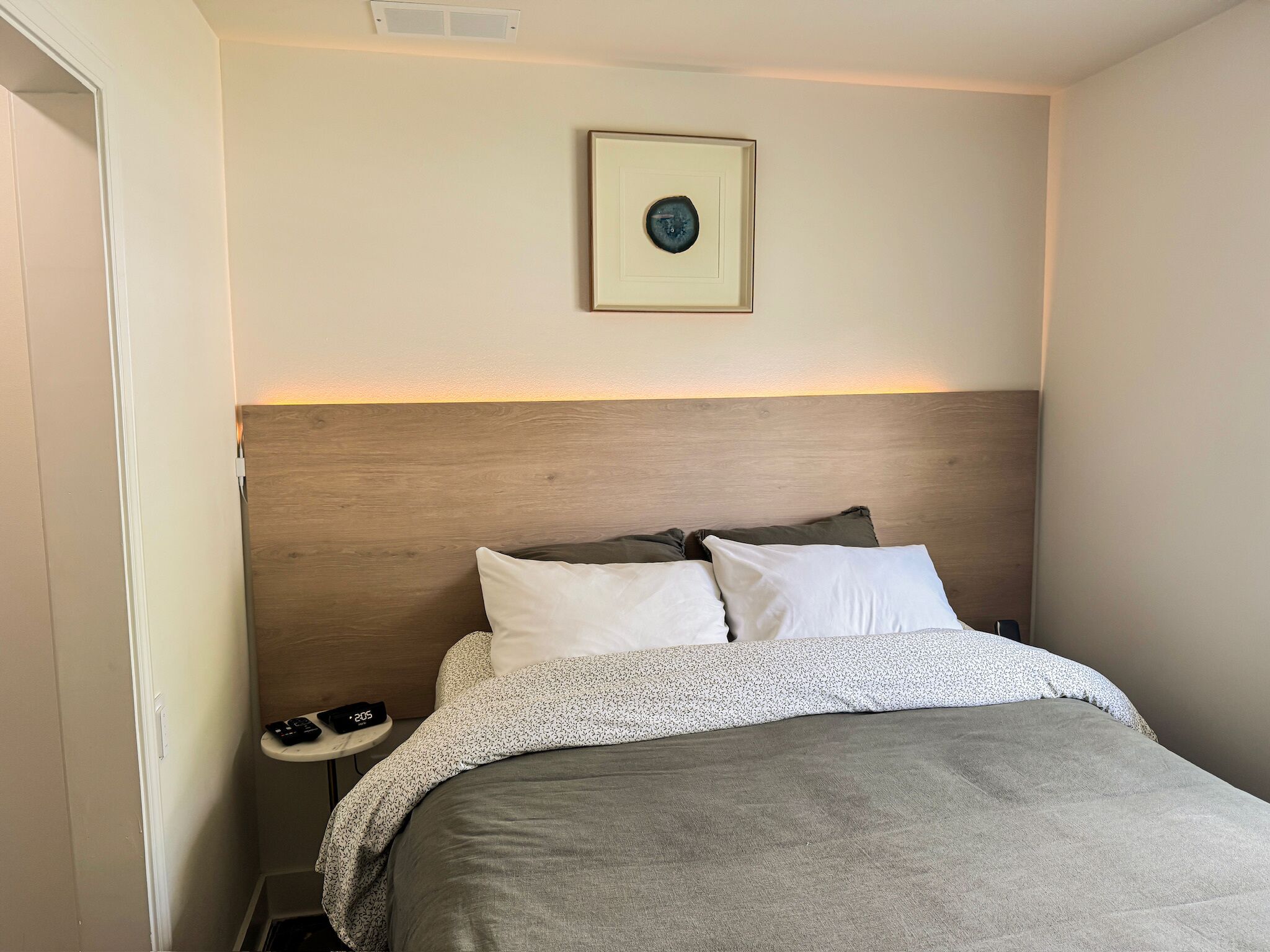 Cama bem arrumada com manta cinza, cabeceira de madeira e mesa de cabeceira com relógio.