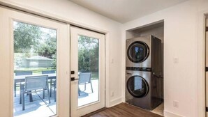 Un cuarto de lavandería con entrada de doble puerta, lavadora y secadora de acero inoxidable, y vista del patio exterior a través de puertas de vidrio.