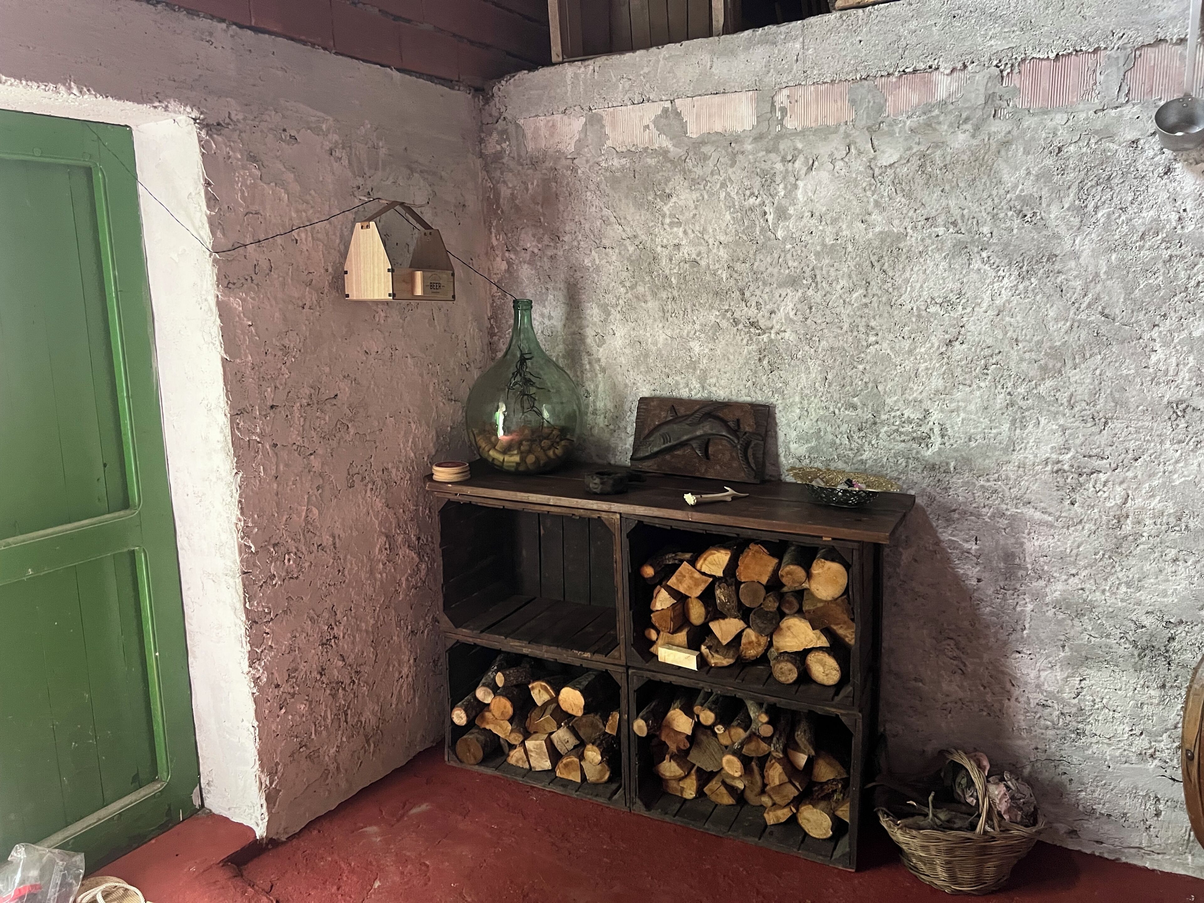 Un coin rustique avec une porte verte, une étagère sur laquelle se trouve une bouteille verte, une sculpture de poisson et un panier de bois de chauffage.