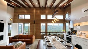 A spacious living area with a long dining table set for six, wooden beams on the ceiling, and large windows offering a view of the forest. - Luxury Alpine Retreat - Hot Tub & Sauna - Sleeps 16 - Cliffhanger Chalet (Comox Valley C (Puntledge-B*)