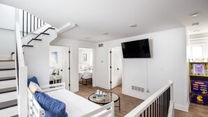 A modern, well-lit room with a staircase, a flat-screen TV mounted on the wall, and a small table with books.