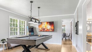 A pool table in a room with wooden flooring, a television mounted on the wall, and a door leading to another room. - Cozy Home w/ Pool Table, Arcade & Firepit (Spartanburg)
