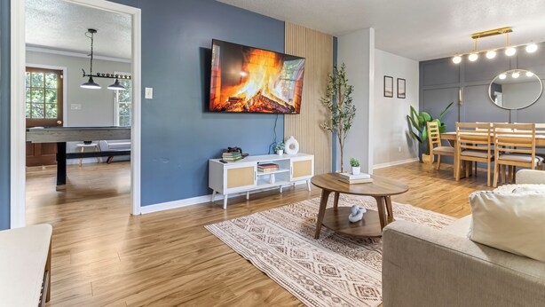 A modern living room with a TV, a dining area, and a kitchen visible in the background. - Cozy Home w/ Pool Table, Arcade & Firepit (Spartanburg)