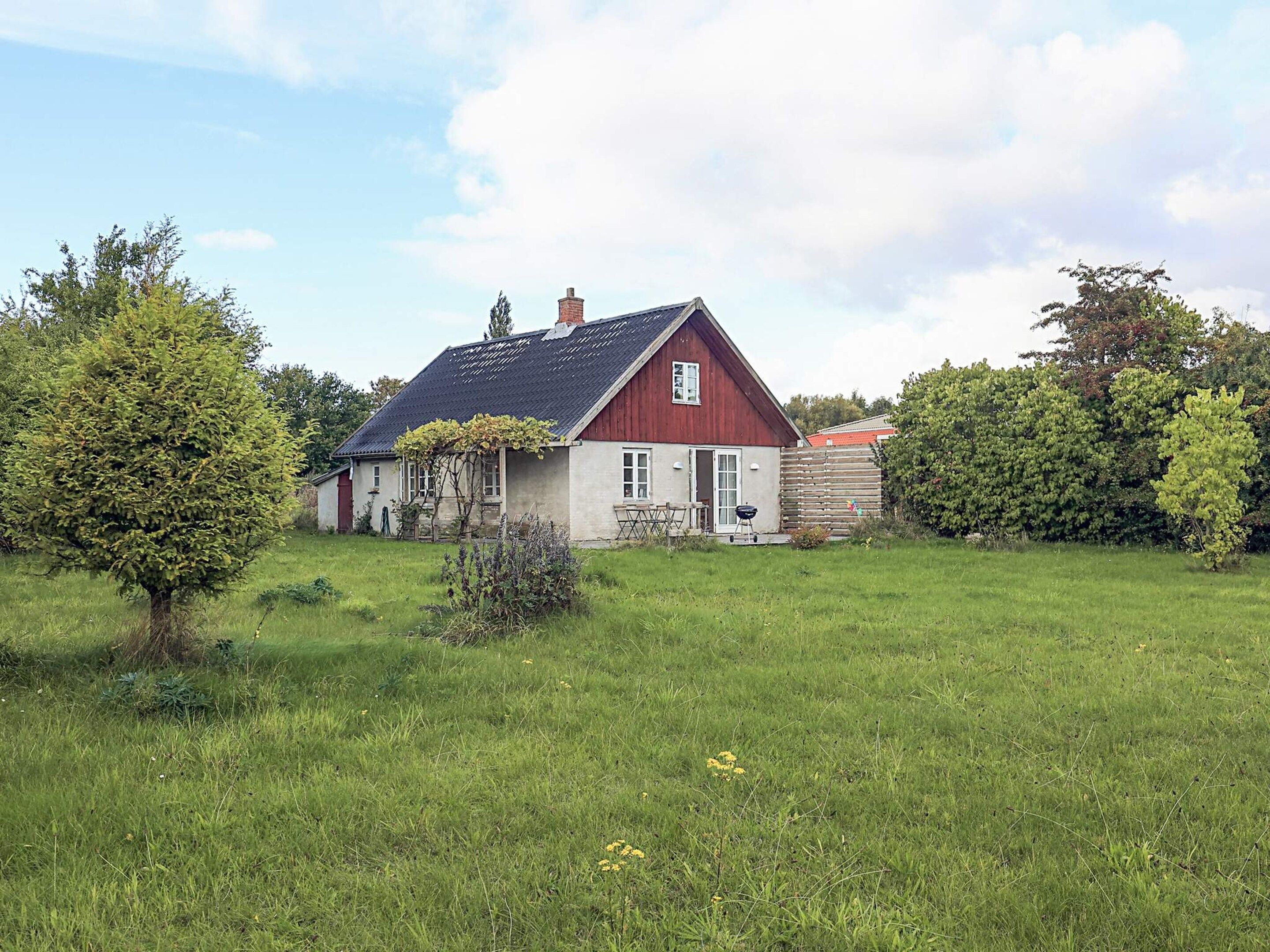 A single-story house with a red exterior and a grey roof, surrounded by a grassy yard and trees.