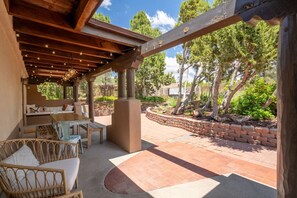 Eine überdachte Terrasse mit Sitzgelegenheiten im Freien, einem Kamin aus Stein und Blick auf einen angelegten Garten mit Bäumen.