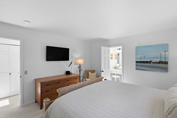 Un dormitorio con una cama, un aparador de madera, un escritorio con una silla, una lámpara, un televisor en la pared y un cuadro.