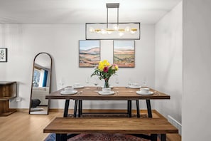 A dining room with a wooden table set for six, a vase with flowers, and two landscape paintings on the wall.