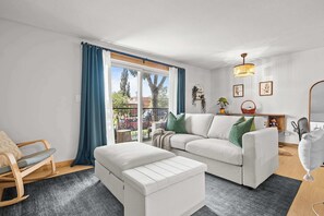 Uma sala de estar moderna com um sofá branco, uma poltrona de balanço de madeira, um banquinho branco e uma janela grande com cortinas azuis.