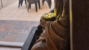 Eine Buddha-Statue blickt auf eine Terrasse mit Tisch, Stühlen, einem Topfpflanzen und einer Tür im Hintergrund.