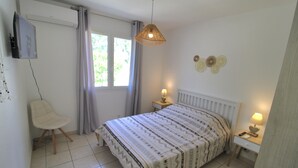 Ein Schlafzimmer mit einem Bett, einem Sessel, einem Nachttisch mit Lampe, einem an der Wand befestigten Fernseher und einem Fenster mit Vorhängen.