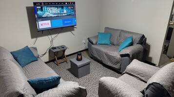 A living room with two grey couches, a small coffee table, and a flat-screen TV mounted on the wall displaying a movie.