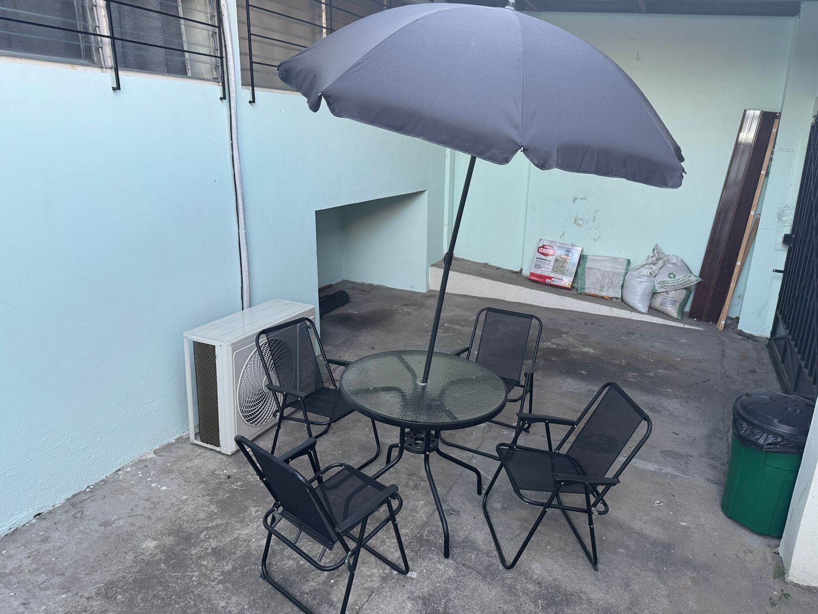 Una terrassa exterior amb una taula rodona, quatre cadires, un parasol gran, un aparell d'aire condicionat i un safataire.