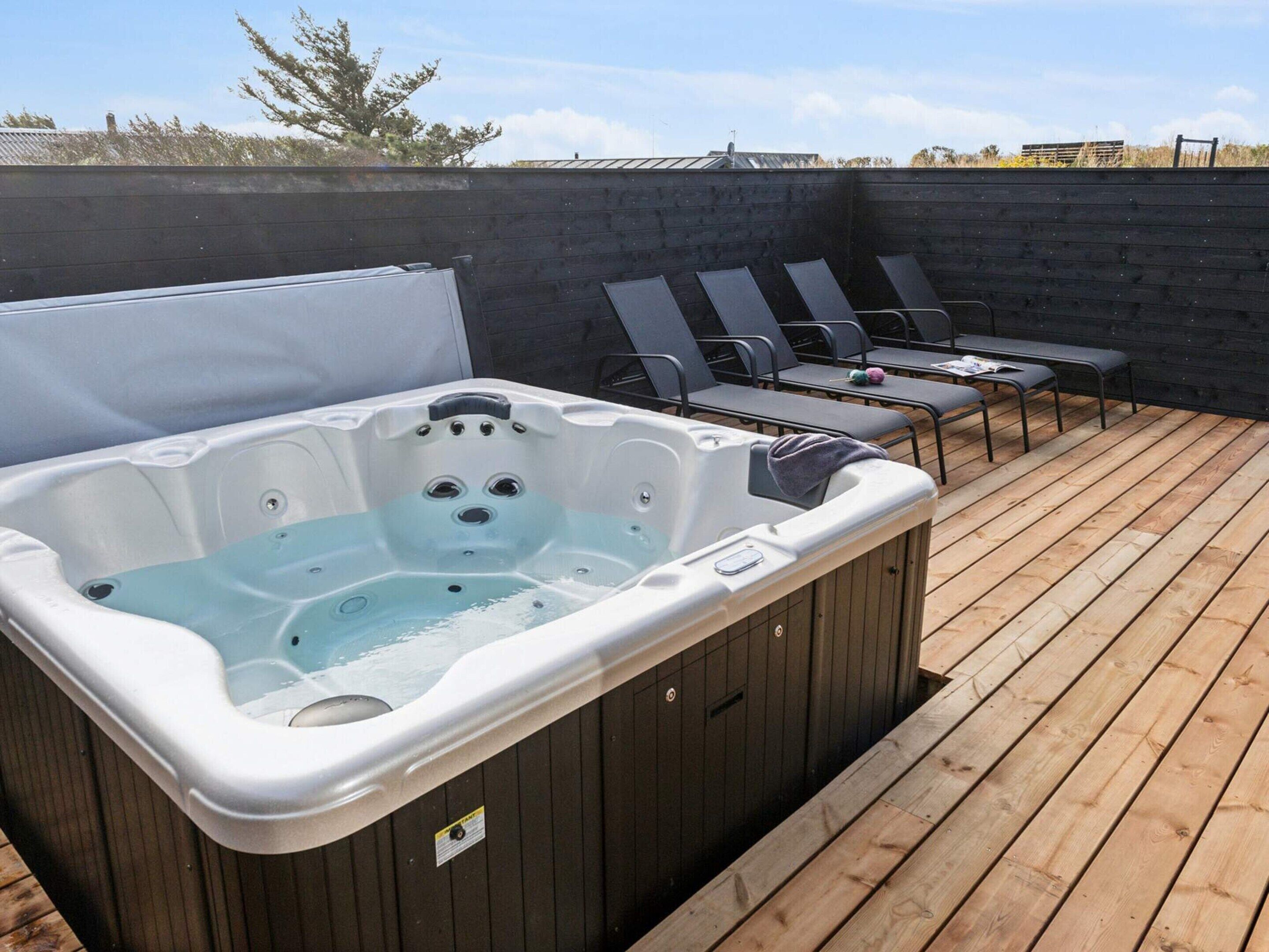 Un jacuzzi en una terraza de madera con sillones reclinables y un muro de madera al fondo.