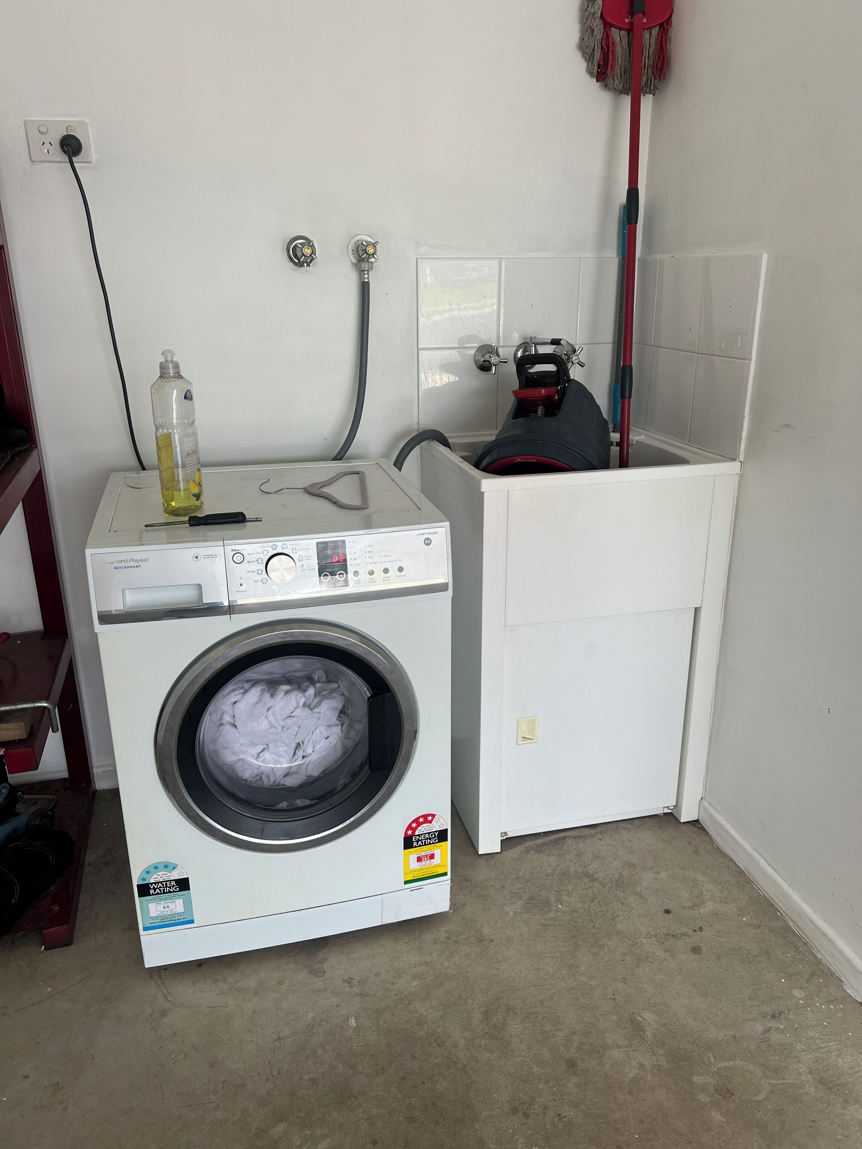 A compact laundry area with a front-loading washing machine, a hose, and a red mop.