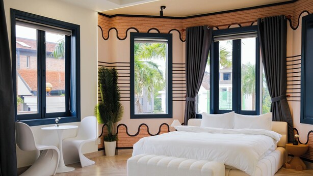 Una habitación de hotel moderna con una cama grande, una mesita redonda pequeña, dos sillas blancas y una planta en maceta.