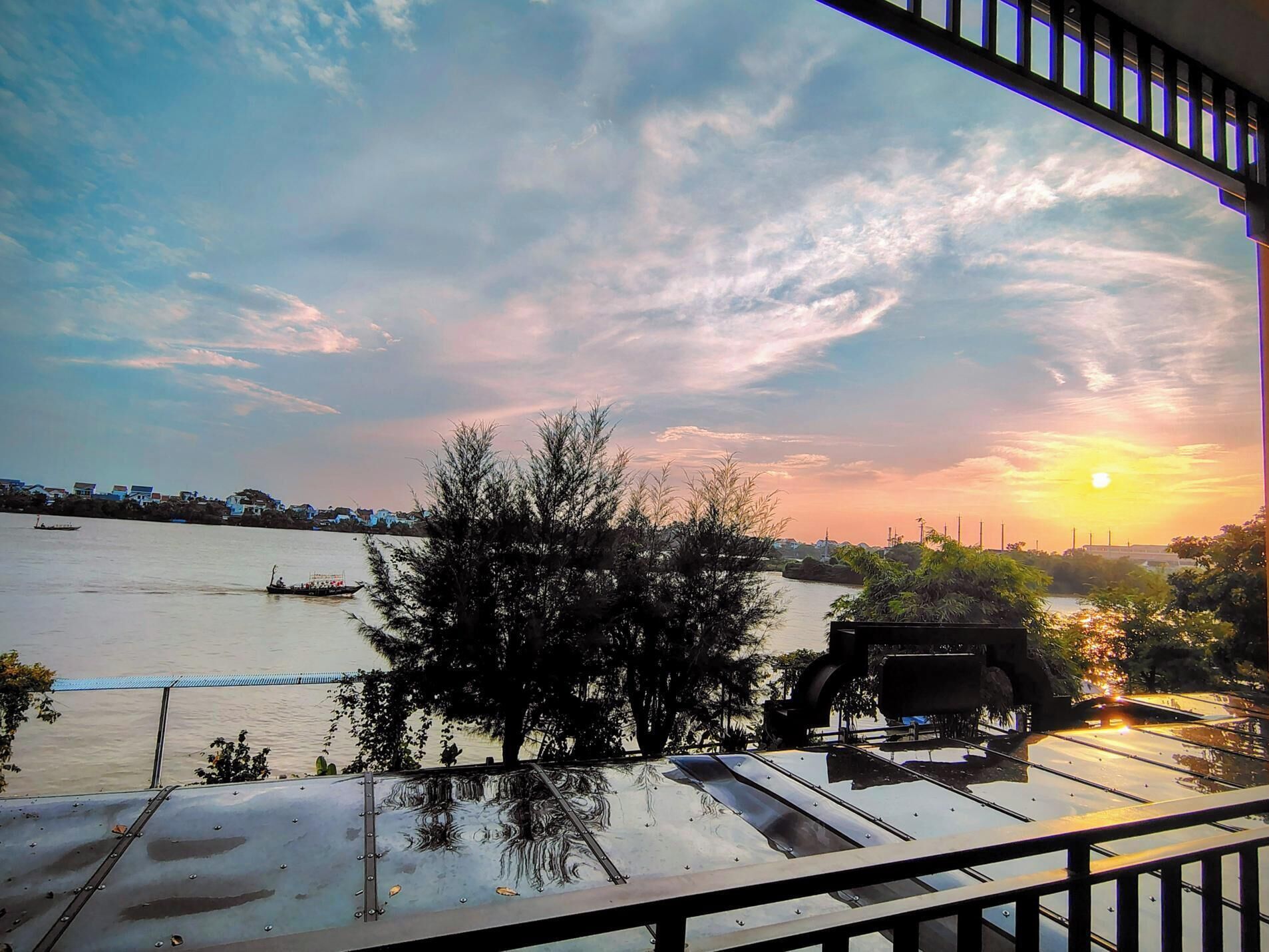 A sunset view from a balcony overlooking a river with boats, trees, and a distant cityscape.
