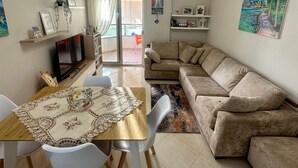 A modern living room with a beige sofa, a dining table with a lace tablecloth, and a TV on a stand.