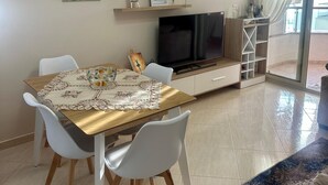 A modern living room with a dining area, featuring a flat-screen TV, a coffee table with a lace table runner, white chairs, and artwork on the walls.