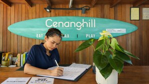 A person writing at a wooden desk with a surfboard-shaped sign behind them that reads "Cenanghill" and "EAT PLAY". - Seaside Cenang (Langkawi)