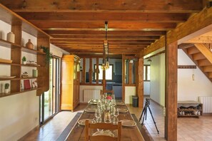 A dining area with a long wooden table set for a meal, wooden beams on the ceiling, and shelves with various items on the left wall.
