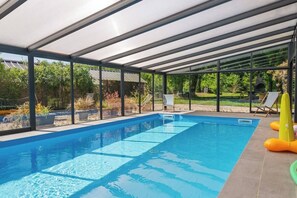 A covered swimming pool with clear glass panels, a tiled deck, and a view of a garden and trees.