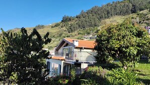 Une maison à deux étages dotée d’un balcon, entourée d’une végétation luxuriante et avec des collines en arrière-plan.